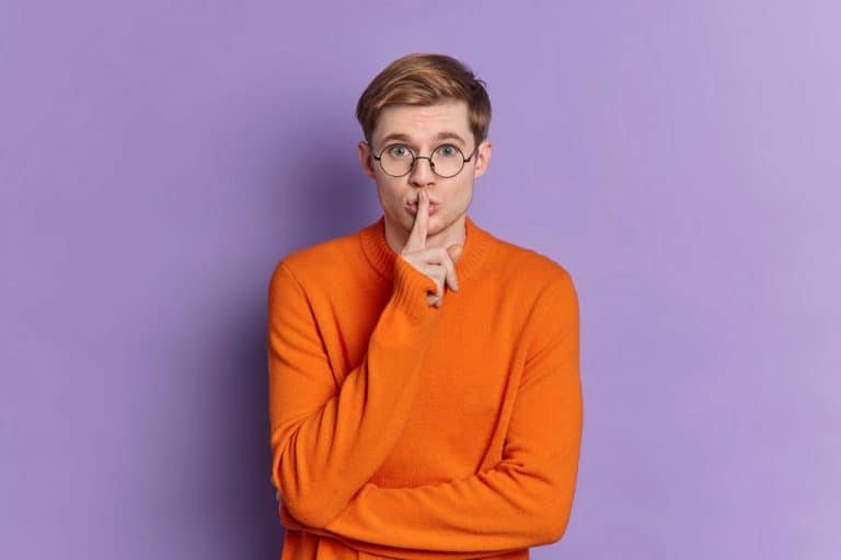 Portrait of teenage boy makes silence gesture keeps fore finger over lips makes shh sign tells secret has mysterious face expression wears bright orange sweater isolated on purple background