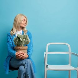 Calm serious blonde woman concentrated on chair holds potted cactus feels lonely lives alone dressed in fashionable clothes isolated over blue background. People retirement lifestyle concept