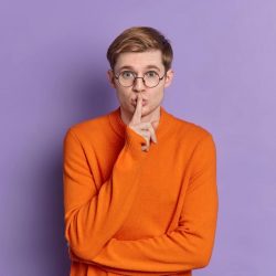 Portrait of teenage boy makes silence gesture keeps fore finger over lips makes shh sign tells secret has mysterious face expression wears bright orange sweater isolated on purple background